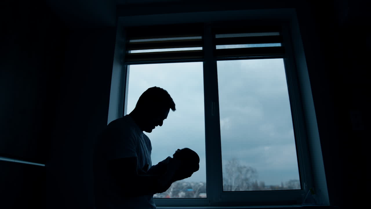 Big strong man's silhouette with a baby in hands. Father holding his newborn standing in the dark room in front of the window.