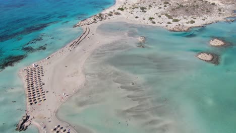 Drone-view-in-Greece-flying-over-Elafonisi-white-sand-narrow-beach,-clear-blue-water-on-the-sides-and-many-umbrellas-on-a-sunny-day