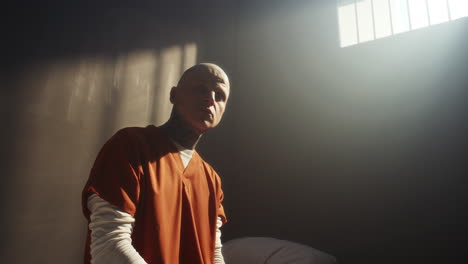 portrait of tattooed inmate sitting in prison cell under barred window