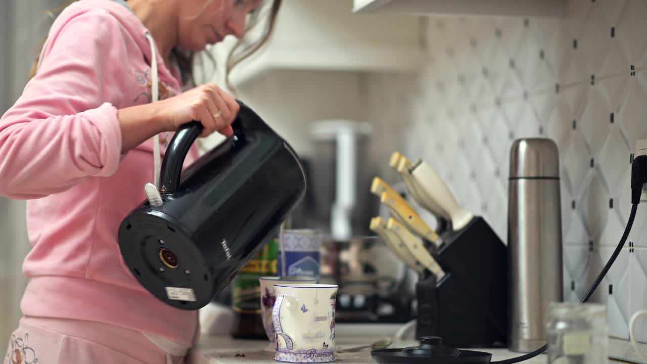 In a warm kitchen, a person pours hot water from a kettle into a cup, preparing tea for a calm start to the day. The space is filled with light and comfort