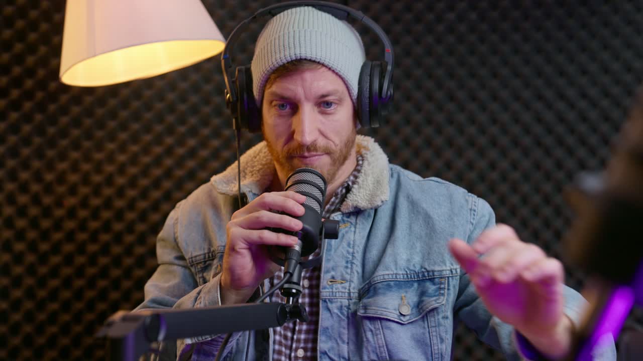 Podcast or radio host looking at camera and waving hello at start of live show. Man put headphones on and setting up microphone in professional recording booth or studio. Beginning of streaming show