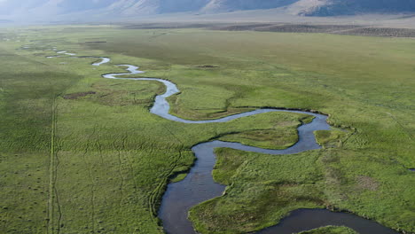 a winding river peacefully meanders through lush, expansive green fields with distant mountain ranges, drone