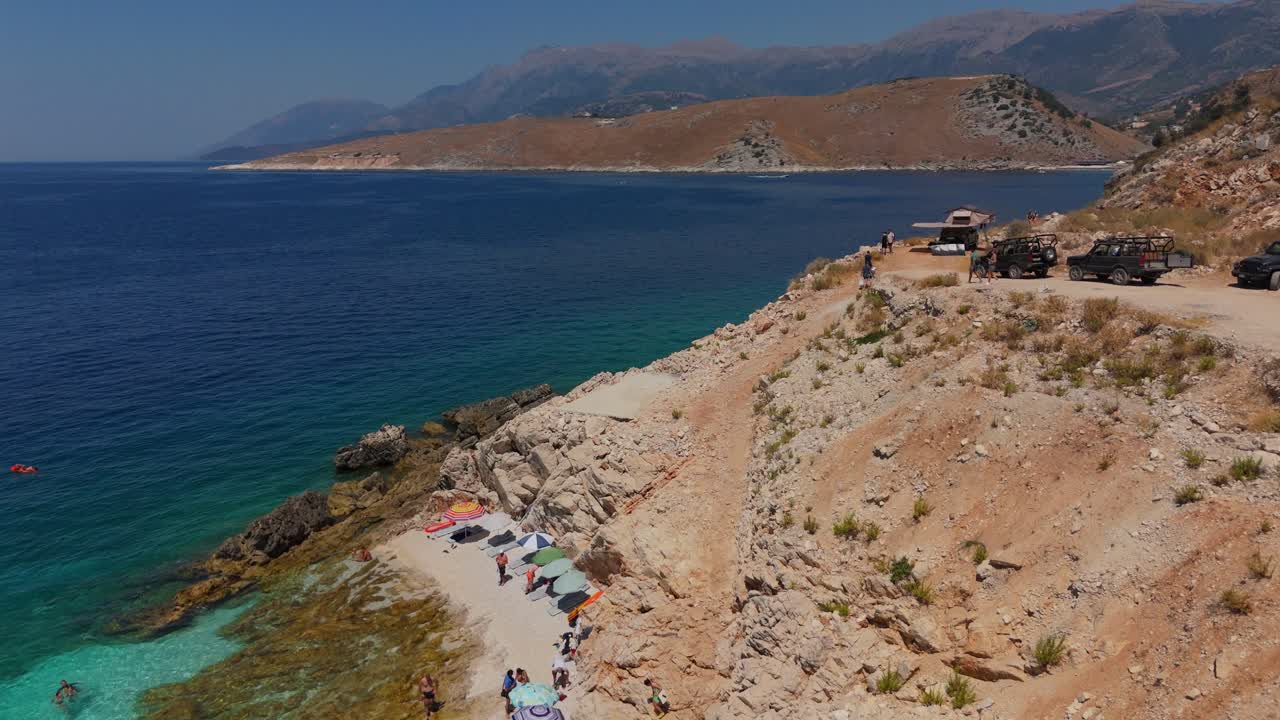 un jeep conduce a lo largo de un camino de tierra costera con una playa turquesa debajo en albania