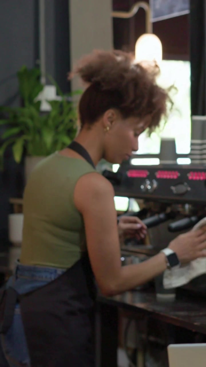 Video verticale: Un barista con maglietta verde e grembiule che impugna il portafiltro, prepara il caffè e pulisce la lattiera.