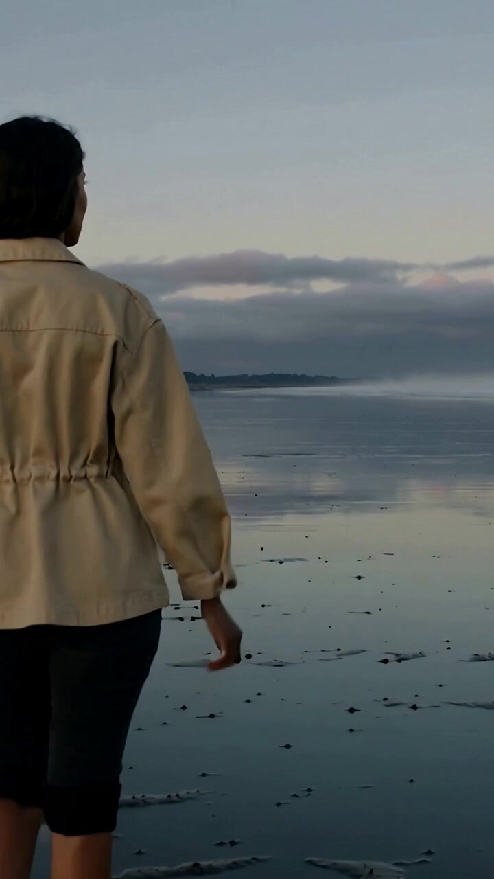Vertical video: Starting woman in tan jacket pants walking along wet sand looking to sea