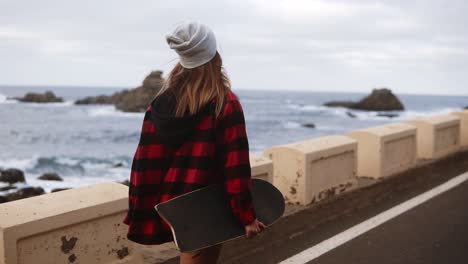 Rare-View-Of-Sporty-Builded-Woman-In-Hat,-Coat-And-White-Sneakers-Enjoying-Time-By-The-Seaside-On-A-Cloudy-Day-At-Sunset,-Walking,-Holding-A-Skateboard,-Slow-Motion