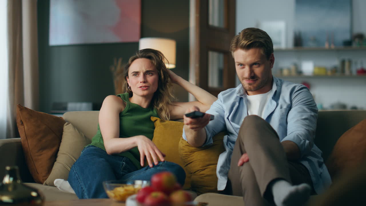 Afslappede partnere slapper af på sofaen og ser tv om aftenen. Smilende mand skifter kanaler.