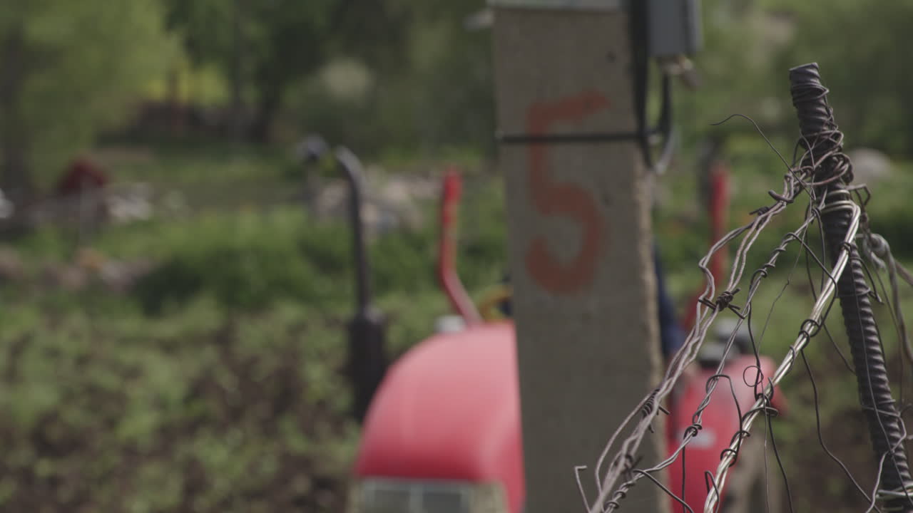 un retrato borroso de un granjero conduciendo un tractor rojo arando el campo visto detrás del poste de hormigón con una cerca de alambre de hierro enredado en una pequeña granja de pueblo en moliti, georgia