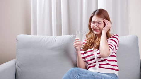 Mujer-Joven-Asiática-Sentada-En-El-Sofá-Bebiendo-Agua-Para-Relajarse-Durante-Un-Fuerte-Dolor-De-Cabeza