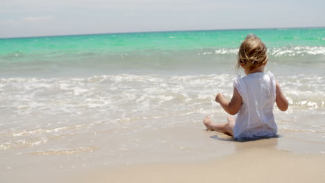 Kleines-Mädchen-Sitzt-An-Einem-Strand-In-Der-Nähe-Des-Wassers