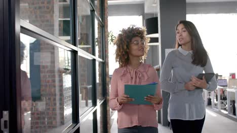 happy biracial female colleagues walking through office talking, with tablet and folder, slow motion