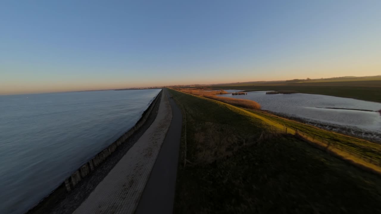 Bicycle path next to a Dutch dyke during sunset with two cyclists in the distance. Aerial follow shot