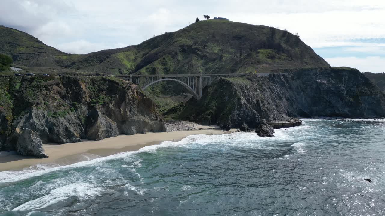 Drone flies over the water showing a beautiful beach beneath one of the iconic bridges along highway 101 near Big Sur, California