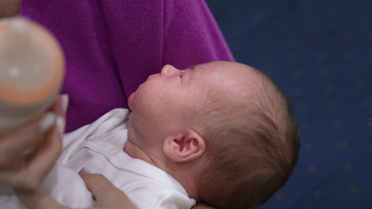Little baby opens its mouth in hunger. Mother gives a bottle to her child, it suckles and milk drops appear on its cheek. Close up.