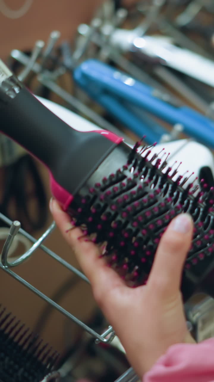 Hand of a female shopper closely examining a hair styling tool in a salon store, the shopper is holding a brush-like hair device, observing its details, with a rack of other hair tools
