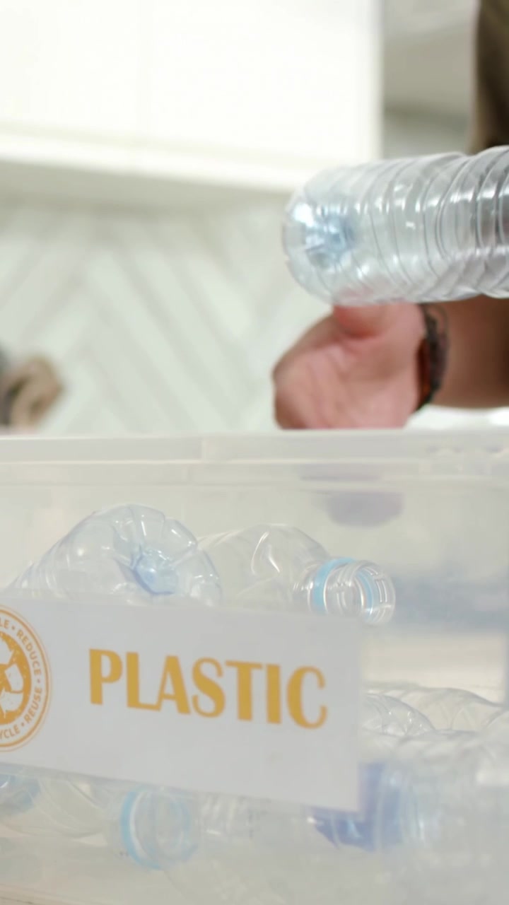 Video verticale: Una donna in rosa individua una vasca di PLASTICA a casa e consegna una bottiglia a un uomo in abiti color oliva per il riciclo.