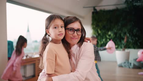 Portrait-of-a-female-teacher-in-a-white-shirt-with-a-bob-hairstyle-and-glasses-hugging-her-student-in-a-club-for-preparing-children-for-school-against-the-backdrop-of-children-having-fun-at-recess