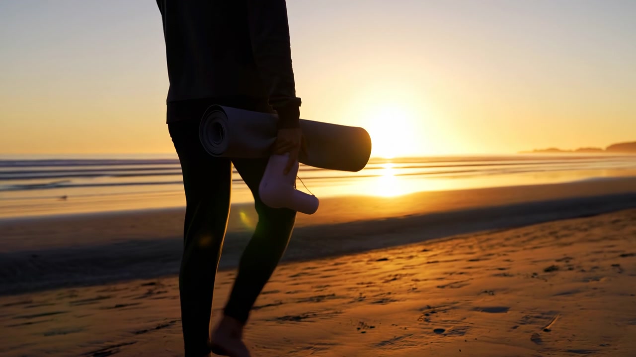 Silhouette of person walking barefoot beside ocean at sunrise, carrying rolled yoga mat and small handheld device, with warm backlight, sandy shoreline, gentle waves, soft reflections, and peaceful