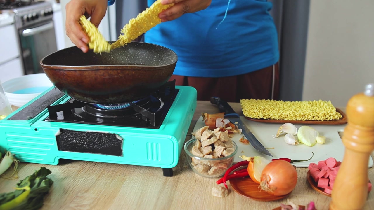 Adegan persiapan makanan yang menampilkan mi kering dijatuhkan ke dalam wajan.