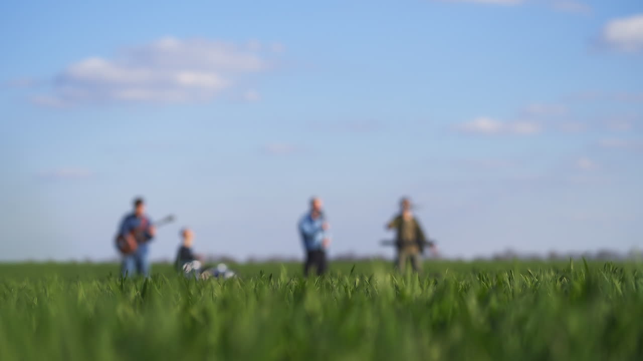 Music band of four guys perform song and instrumental music for drone footage. Gradual view transition from men at backdrop to green grass at foreground.