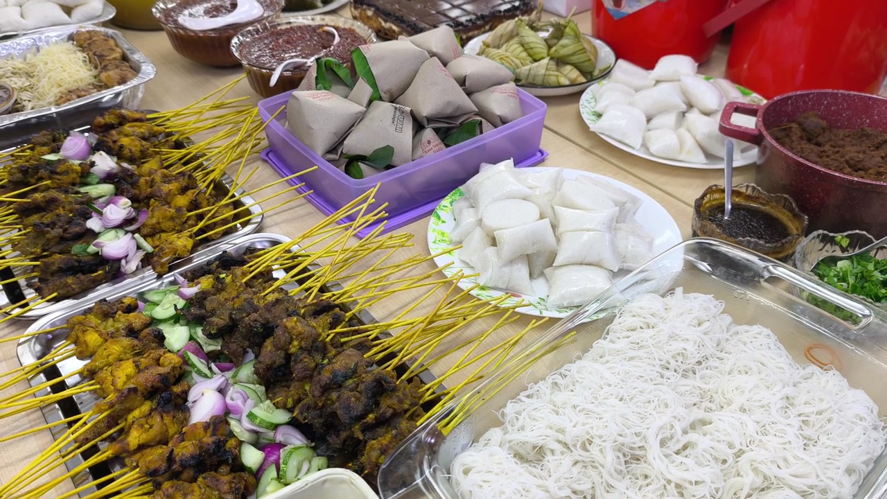 Pesta Hari Raya yang meriah menampilkan hidangan tradisional Malaysia yang dipajang. Makanan yang disajikan meliputi ketupat, saus kacang, dan pilihan sate. Para tamu merayakan Idul Fitri dengan cita rasa lokal yang kaya.