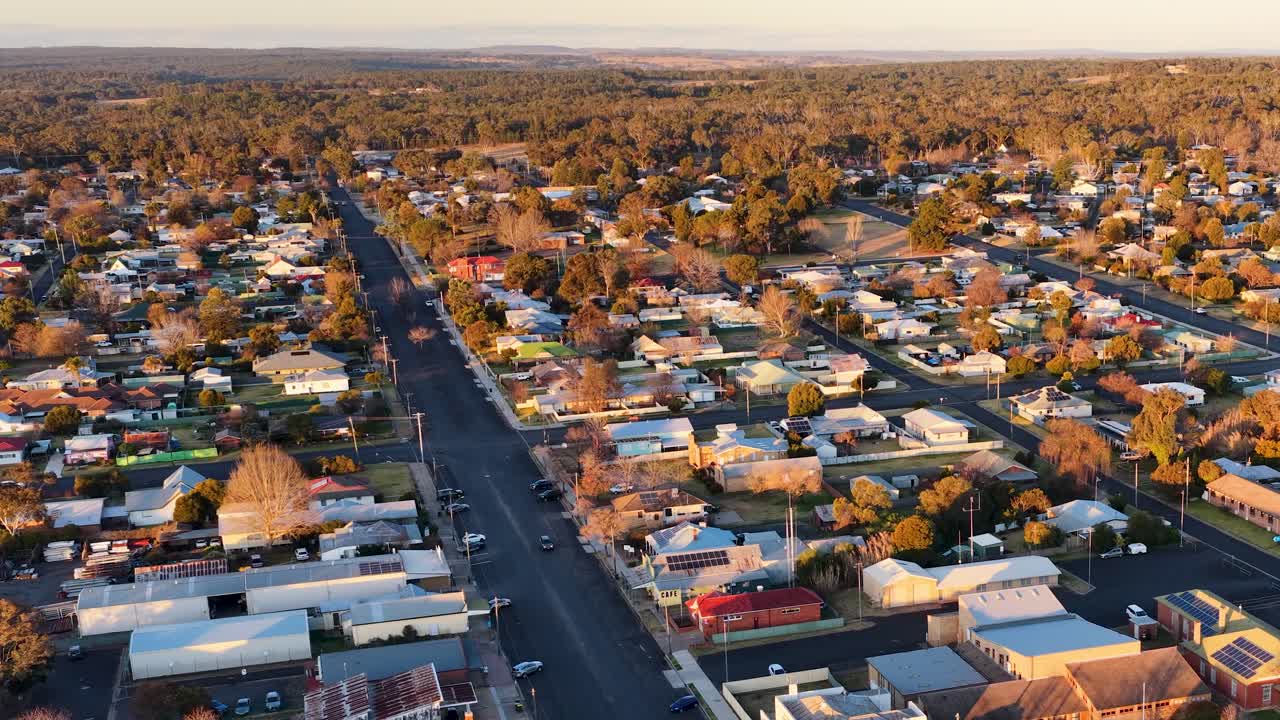 Drone camera glides above Coonabarabran, New South Wales, revealing sunlit rooftops, tree-lined streets, and a tranquil small-town atmosphere during golden hour