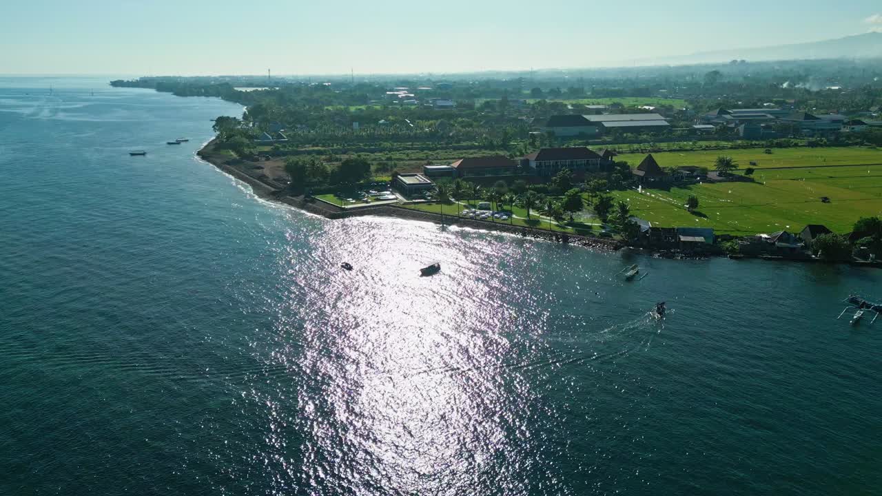 Drone aerial footage of North Bali coastline showing gentle waves, traditional fishing boats, village houses, and lush tropical trees under bright daylight in a popular coastal tourist area