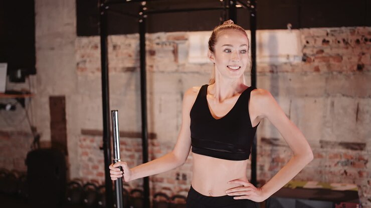 cámara lenta de una joven deportista sonriente sosteniendo una barra mientras está de pie con la mano en la cadera en el gimnasio