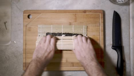 lapso de tiempo de la vista de arriba hacia abajo en dos manos en una tableta de corte de madera en el mostrador de la cocina rodando ingredientes en una hoja de alga nori y estera haciendo un rollo de sushi