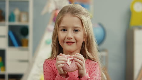 portrait of the beautiful sweet blonde little girl smiling to the camera with a pink donut in hands in her cozy room