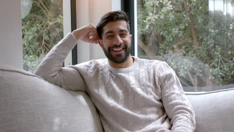 Portrait-of-happy-biracial-man-sitting-on-sofa-smiling-at-home,-slow-motion