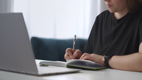 teen-girl-is-doing-homework-in-notebook-using-laptop-for-looking-for-information-learning-in-home-after-class-detail-view-on-table