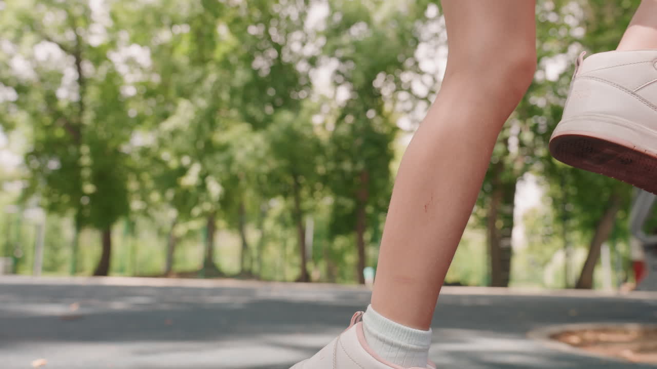 Primo piano di una figlia bianca che salta lungo il sentiero del parco, scarpe da ginnastica bianche e calzini alla caviglia, cadenza costante e movimento ritmico, ombra a chiazze degli alberi e calore estivo, passi giocosi e sicuri.