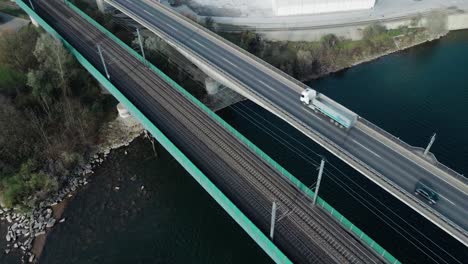 a passenger train crossing a river via a railroad bridge located next to a highway where trucks drive next to it