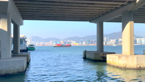 Stunning-View-of-Victoria-Harbour,-Hong-Kong:-Cargo-Ship-Sails-as-North-Point-Pier-Comes-to-Life-with-Ships-and-Boats,-Cityscape,-Waterfront,-Travel