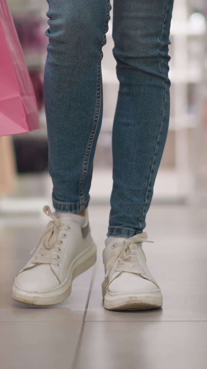 Leg view of shopper walking confidently through retail clothing store aisle carrying vibrant pink and green shopping bags, in a modern, neatly arranged retail environment