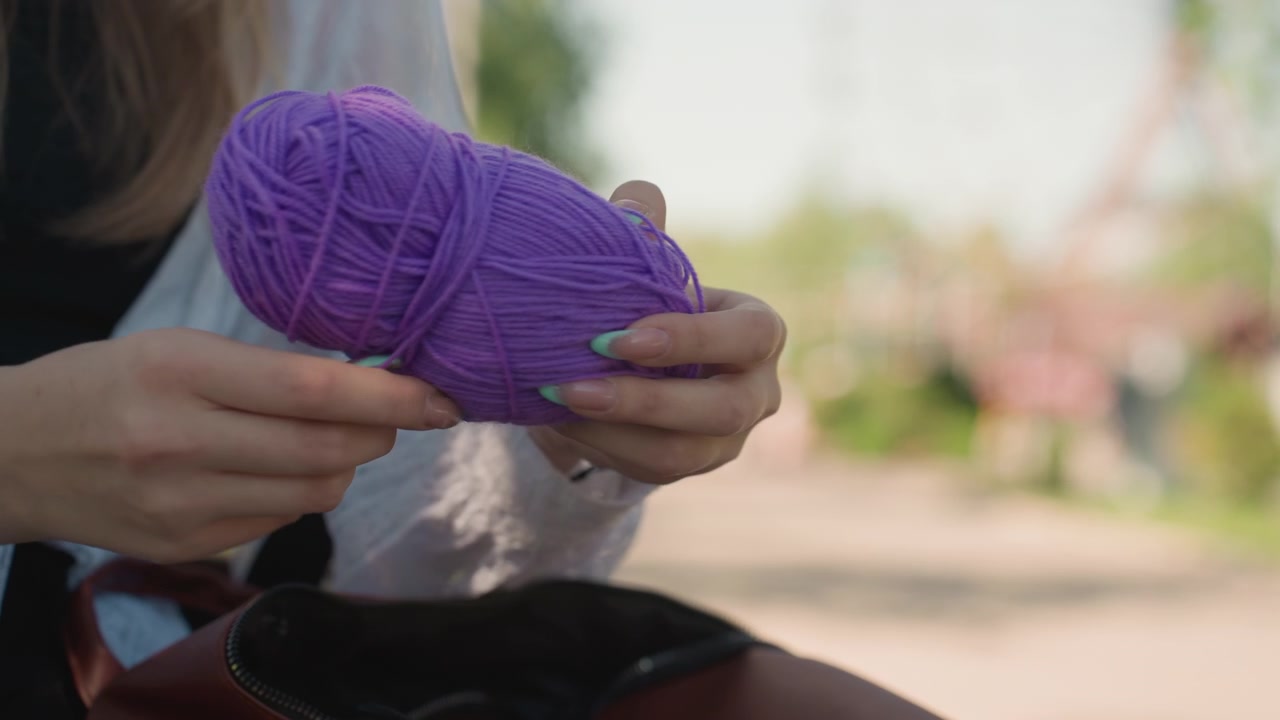 Primer plano de una mujer joven preparándose para tejer con hilo morado en un entorno al aire libre. La mujer sostiene una bola de hilo y una aguja de tejer, concentrándose en sus manos mientras desenreda o ajusta los materiales.