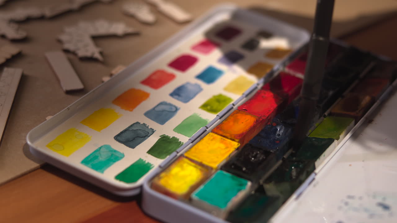 Brush Dipping Into Watercolor Pans, Wet Tip Pulling Deep Pigment From Red And Blue Wells, Stencils And Paper. Media