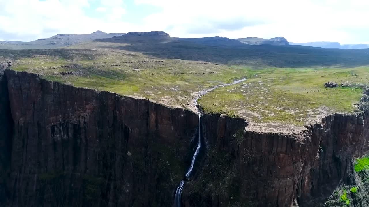 Aerial drone footage of Tugela Falls waterfall in South Africa, where a narrow river is falling off a steep brown cliff. The top has green nature and grass growing, mountains visible in the background