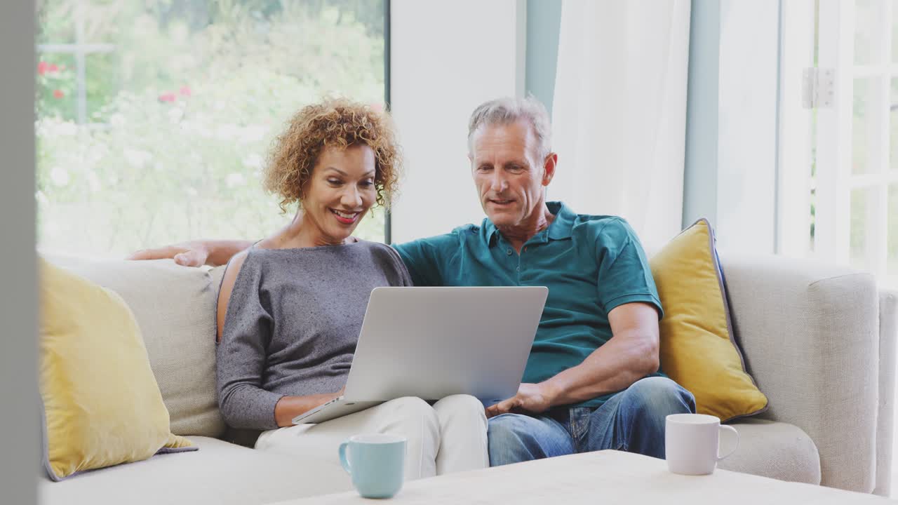 Senior Retired Couple Sitting On Sofa At Home Shopping Or Booking Holiday On Laptop