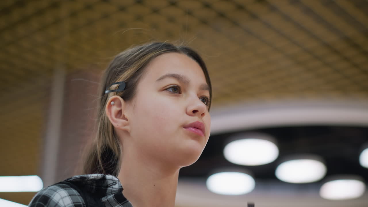 Casual young girl walks in a modern shopping mall, holding a juice cup with both hands, she sips slowly while looking up at the ceiling, then gently lowers her head, surrounded by bright interior