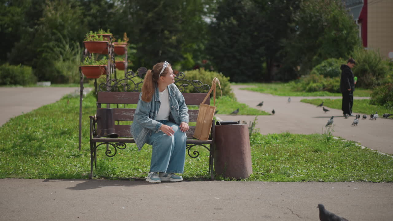 Ung kvinde sidder på bænk, afslappet studerende tager eftertænksom pause blandt duer og grønt, denim-outfit, stofpose på sædet, solrig forstadsparksti, rolig middagsstemning og oprigtigt portræt