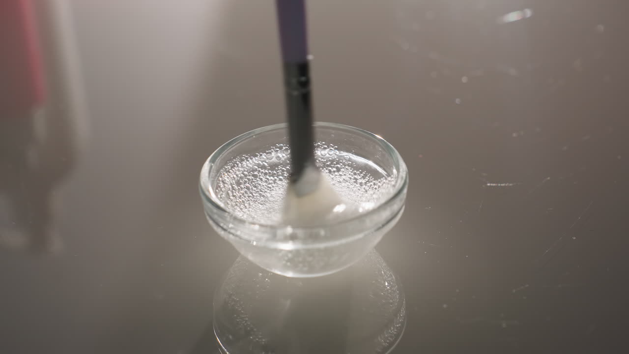 Gloved hands stir bubbly gel with soft facial brush in clear glass bowl on reflective surface, highlighting hygienic spa prep, soothing texture and expert skincare blending ritual