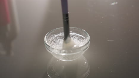 gloved hands stir bubbly gel with soft facial brush in clear glass bowl on reflective surface, highlighting hygienic spa prep, soothing texture and expert skincare blending ritual