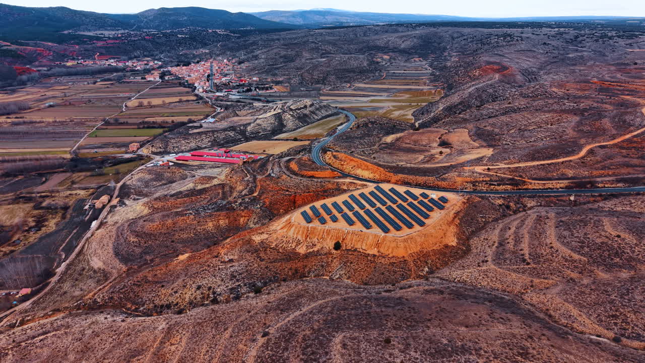 Wide aerial view of solar installation and rural village. Long distance drone shot showing photovoltaic array, hills, winding road and small town in background
