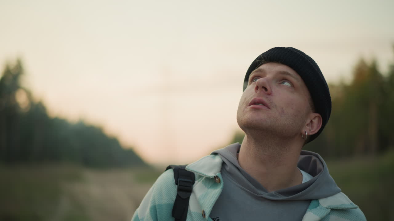 Primer plano de un mochilero caucásico fumando al atardecer con la mano cerca de la boca, escudriñando el cielo, con las correas de la mochila visibles, línea de árboles distante, suave resplandor del atardecer, expresión introspectiva, ambiente de planificación de la ruta, cinematográfico, superficial