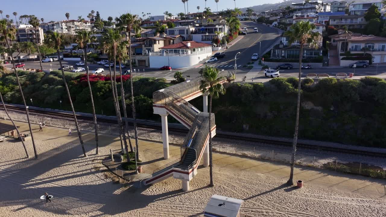 Ripresa aerea orbitante con drone del ponte T-Street che attraversa i binari della ferrovia accanto alla spiaggia sabbiosa a San Clemente, California, con palme, case costiere e la luce del mattino lungo la costa