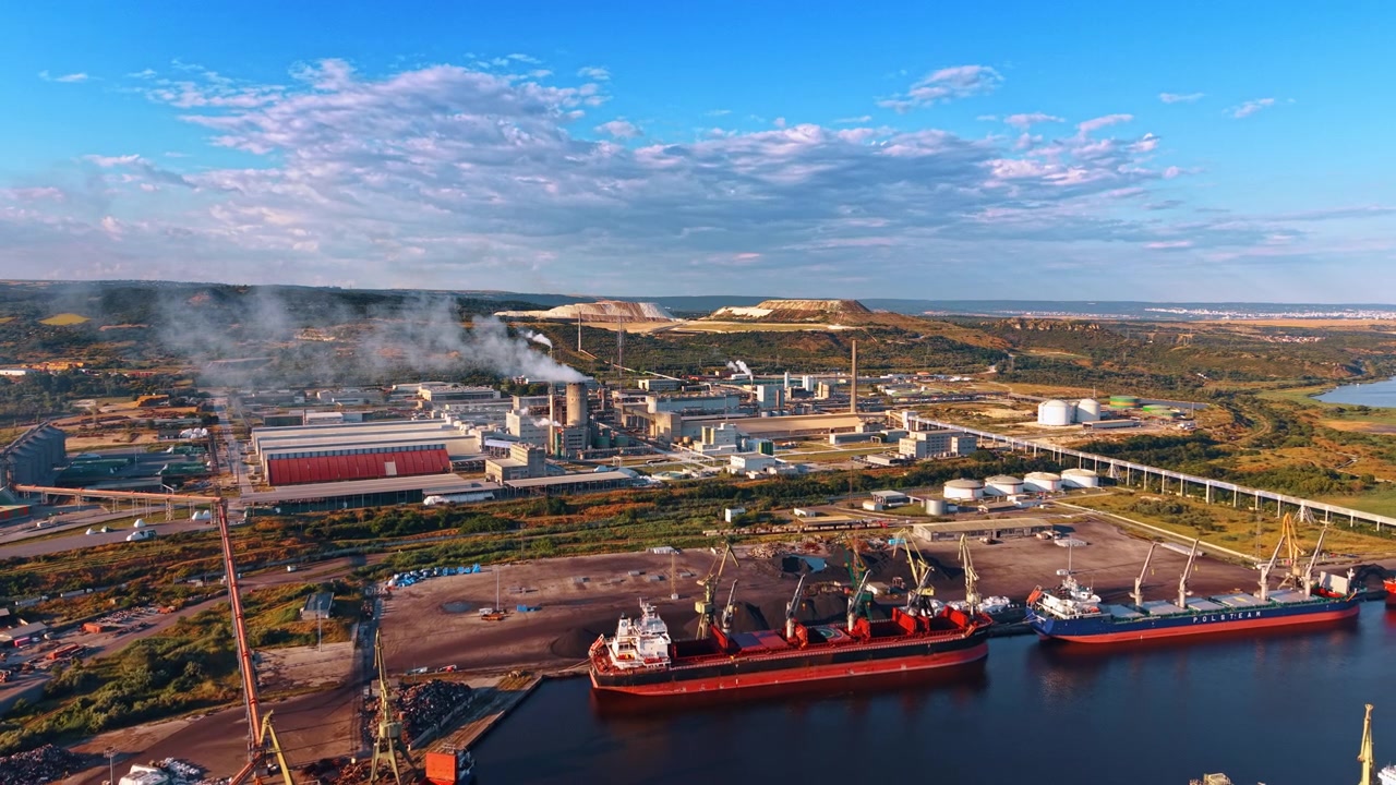 Varna, Bulgaristan, 25 Haziran 2026: Bir fabrikanın yanında endüstriyel bir limana yanaşmış kargo gemileri. Havadan panorama, iki dökme yük gemisini, yükleme vinçlerini, depolama tanklarını ve mavi gökyüzü altında yükselen buharı gösteriyor.