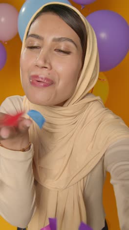 Vertikales-Videostudio-Porträt-Einer-Frau-Mit-Hijab,-Die-Geburtstag-Feiert-Und-Papierkonfetti-Bläst,-Umgeben-Von-Luftballons