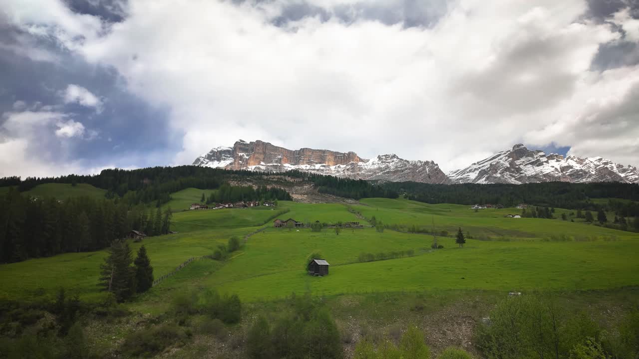 View of a quiet valley in the Dolomites with rolling green hills, a wooden hut, forests, and snowy mountain ridges under a shifting sky